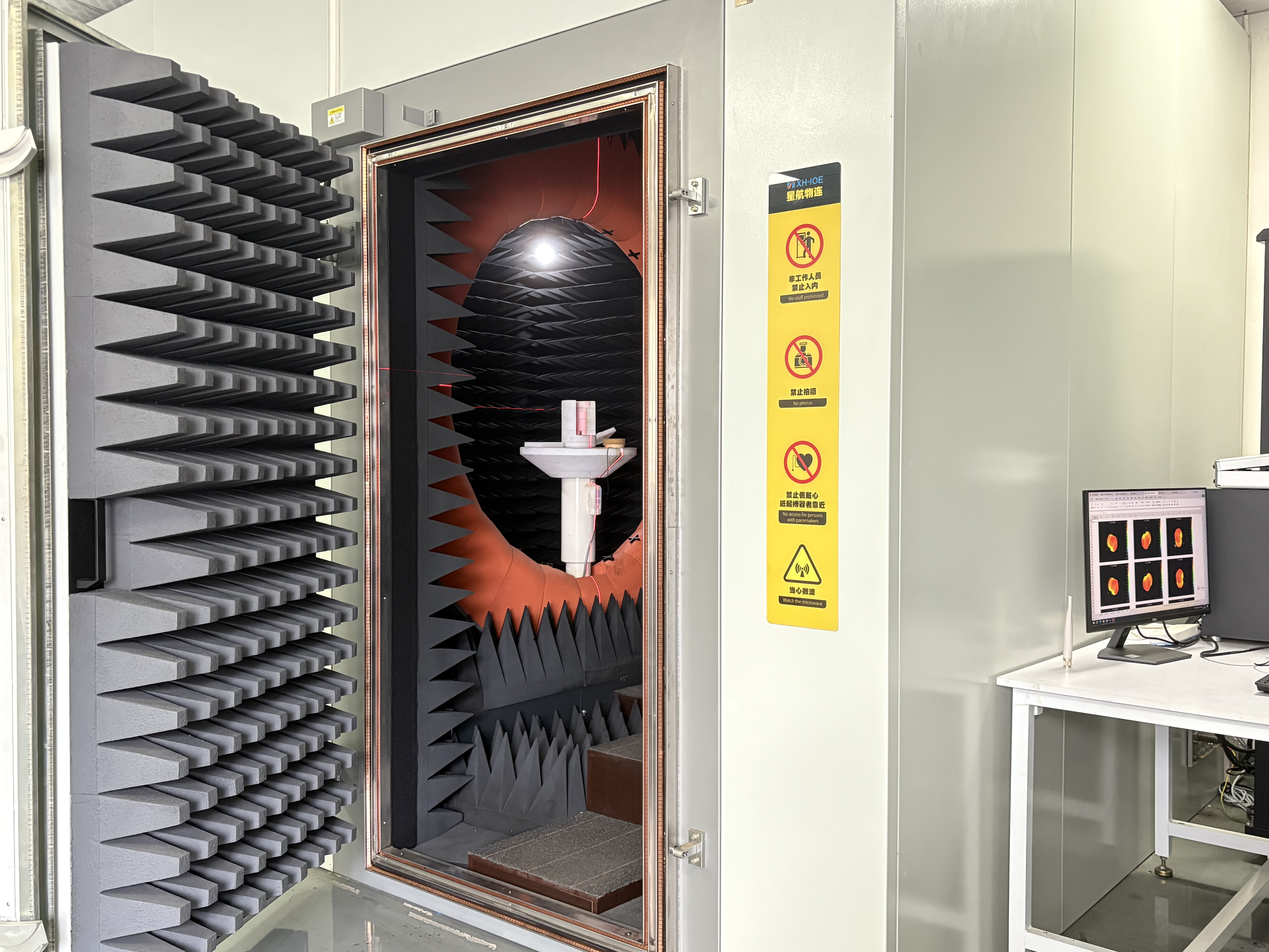 An anechoic chamber with its large, metal door open, revealing the inside lined with pyramid-shaped foam panels. A white device is mounted on a pedestal in the center. A monitor displaying test results sits on a desk to the right, and a yellow warning sign is attached to the exterior wall.